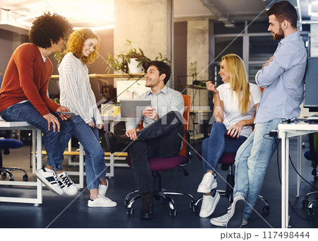 A group of four young professionals engage in a lively discussion in a brightly lit office space. One man sits on a chair holding a tablet, while the others stand around him, conversing and gesturing 117498444