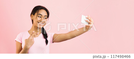 African American girl is standing in front of a pink background, holding a smartphone and posing for a selfie. She looks into the camera with a smile as she captures the moment. 117498466