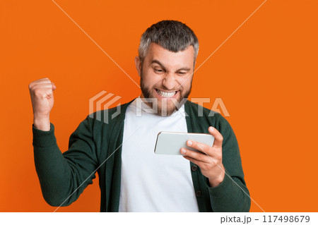 A man in casual clothing holding a smartphone in one hand and raising his fist in a celebratory gesture. The man appears focused and confident. A man in casual clothing holding a smartphone in one hand and raising his fist in a celebratory gesture. The man appears focused and confident. 117498679