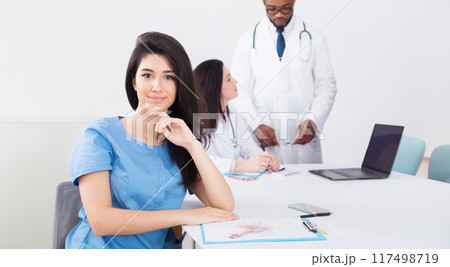 Meeting of medical staff. Nurse smiling to camera at meeting with medical team on background Meeting of medical staff. Nurse smiling to camera at meeting with medical team on background 117498719