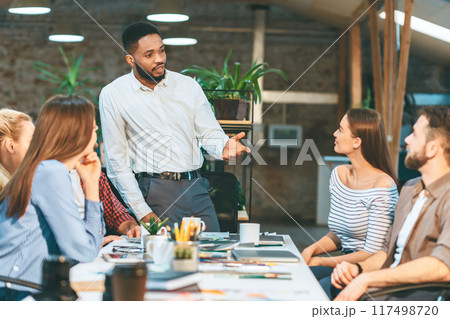 A man stands confidently in front of a diverse group of individuals, engaging in a discussion or presentation. A man stands confidently in front of a diverse group of individuals, engaging in a discussion or presentation. 117498720