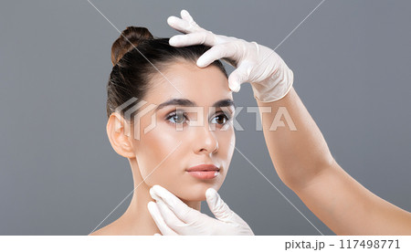 A dermatologist gently examines a young woman face with gloved hands, checking her skin for any irregularities or conditions in a clinical setting. 117498771