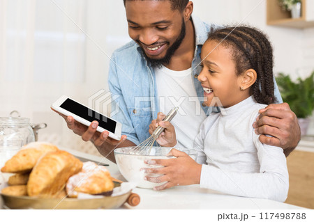 African american father sharing a moment with his daughter, showing something on a tablet in the kitchen, representing family and technology African american father sharing a moment with his daughter, showing something on a tablet in the kitchen, representing family and technology 117498788