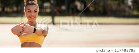 Portrait of sporty African American woman in wireless earbuds warming up before jogging at park, stretching her arms muscles outdoors, feeling healthy and motivated, banner, panorama, free copy space 117498798