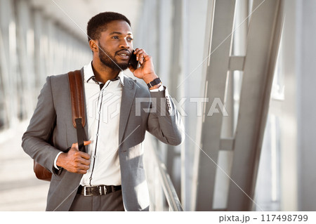 Black man with luggage using his phone outside the airport terminal. The scene is vibrant and professional, highlighting the multitasking nature of business travel. Black man with luggage using his phone outside the airport terminal. The scene is vibrant and professional, highlighting the multitasking nature of business travel. 117498799