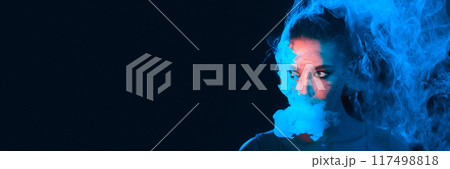 A woman stands partially obscured as blue smoke swirls around her face. The scene appears to be dimly lit, emphasizing the mysterious ambiance, panorama with copy space 117498818