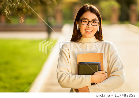 Asian young woman wearing glasses and earbuds is seen walking on a college campus, holding several books and smiling warmly during a sunny day. Asian young woman wearing glasses and earbuds is seen walking on a college campus, holding several books and smiling warmly during a sunny day. 117498858