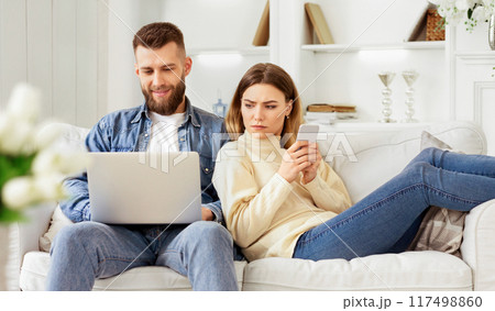 A couple sits on a white sofa in a bright living room, engaged with a laptop and smartphone, respectively, sharing a relaxed moment together. 117498860