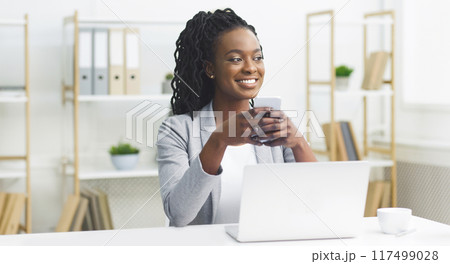 Joyful African American businesswoman at her desk, working with a laptop and a smartphone in a chic office setting. 117499028