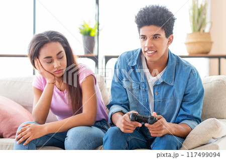 African American teen girl sits on a couch with her head resting on her hand, looking bored. Next to her, a teen guy plays a video game with a controller in his hands, smiling and engaged. African American teen girl sits on a couch with her head resting on her hand, looking bored. Next to her, a teen guy plays a video game with a controller in his hands, smiling and engaged. 117499094