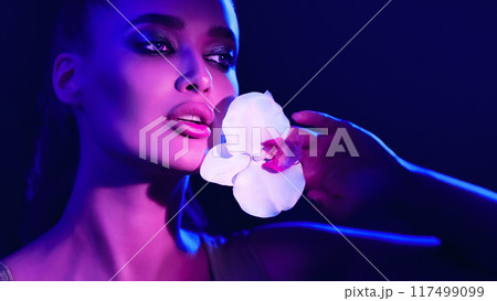 A woman is standing while holding a delicate flower in her hand. She appears focused on the bloom, showcasing its intricate details. 117499099