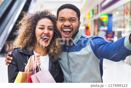 It is fun time. Happy african couple making selfie in mall with paper bags after shopping, panorama It is fun time. Happy african couple making selfie in mall with paper bags after shopping, panorama 117499122