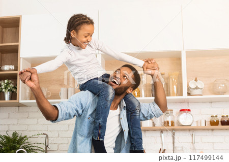 A playful black father lifts his daughter in the air in a well-lit kitchen, encapsulating the joy of african american family and fatherhood A playful black father lifts his daughter in the air in a well-lit kitchen, encapsulating the joy of african american family and fatherhood 117499144