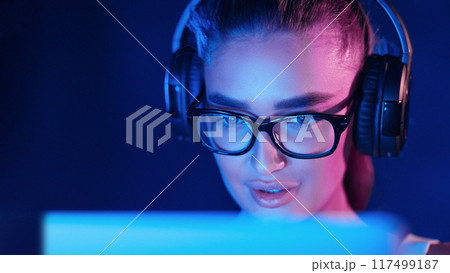 A woman with glasses and headphones is actively engaged in using a laptop. She has a focused expression, typing on the keyboard with the screen illuminated. 117499187