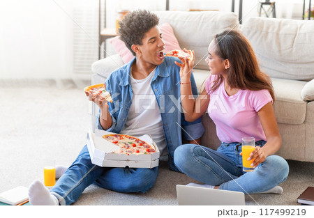 Happy teen couple sits on the floor of their living room, enjoying pizza and drinks. They are both smiling and laughing as they share a pizza. Happy teen couple sits on the floor of their living room, enjoying pizza and drinks. They are both smiling and laughing as they share a pizza. 117499219