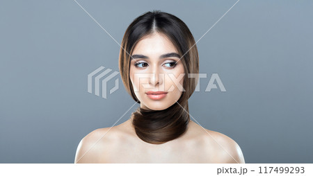 A young woman with long, smooth brown hair wrapped stylishly around her neck poses against a plain gray backdrop. Her expression is serene and thoughtful. A young woman with long, smooth brown hair wrapped stylishly around her neck poses against a plain gray backdrop. Her expression is serene and thoughtful. 117499293