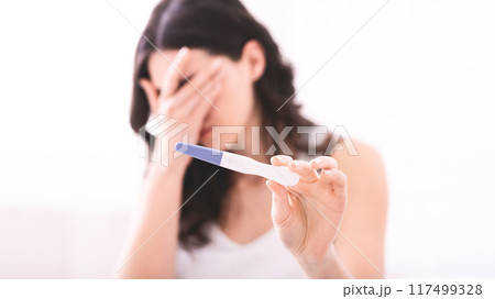 A young woman is holding a pregnancy test in her hand while covering her face with her other hand. She appears to be emotional, possibly in shock or disbelief. 117499328
