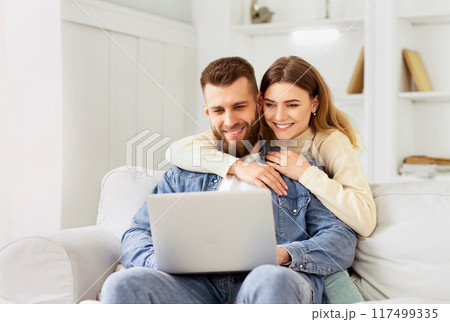Smiling couple sits on the couch, looking at a laptop in a cozy home setting, showing affection and joyful companionship. Smiling couple sits on the couch, looking at a laptop in a cozy home setting, showing affection and joyful companionship. 117499335