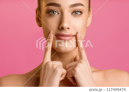 Natural beauty. Young woman applying lip balm, holding fingers near lips, pink background Natural beauty. Young woman applying lip balm, holding fingers near lips, pink background 117499474