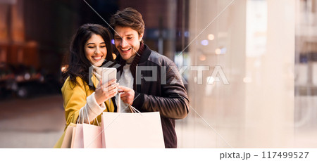 Happy couple ordering taxi on cellphone, walking near city center after shopping on weekend Happy couple ordering taxi on cellphone, walking near city center after shopping on weekend 117499527