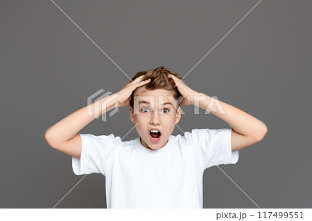 A shocked teen boy is captured with wide eyes and hands clutching his head against an isolated grey backdrop, suggesting alarm A shocked teen boy is captured with wide eyes and hands clutching his head against an isolated grey backdrop, suggesting alarm 117499551