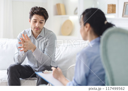 Unhappy man sitting on a couch engaged in conversation with a therapist in a office setting. The man appears focused and attentive while the therapist listens and provides guidance. 117499562