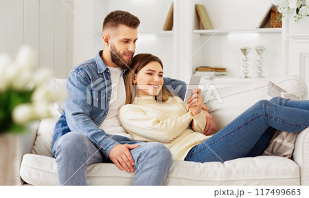 A man and woman are seated on a couch, both focused on a tablet device in front of them. 117499663