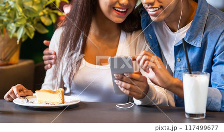 A young black couple is seated at a cafe table, smiling and listening to music through a shared set of earphones, looking at a smartphone together, cropped 117499677