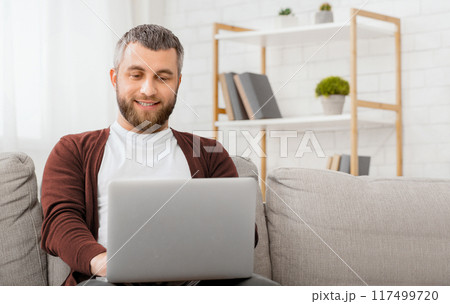 A man is seated comfortably on a couch, engrossed in his laptop screen. He appears focused as he types and navigates the digital device. 117499720