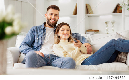 A man and a woman are seated on a couch in a living room setting. 117499732