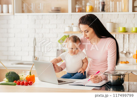 A mother multitasks in a bright, modern kitchen, working on her laptop and taking notes while holding her baby. Fresh vegetables and a pot on the stove are visible in the background. 117499799