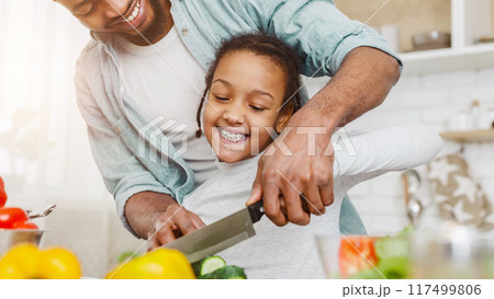 Black cheerful father and cute dughter making salad, cutting cucumber together, cropped 117499806