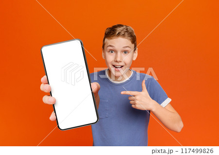 Check it out. Emotional happy boy pointing at blank cell phone screen, orange background with empty space Check it out. Emotional happy boy pointing at blank cell phone screen, orange background with empty space 117499826