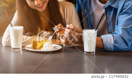 Cropped of guy and girl are seated at a table in a restaurant, enjoying slices of cake.They are both smiling as they take a bite of the delicious dessert. Cropped of guy and girl are seated at a table in a restaurant, enjoying slices of cake.They are both smiling as they take a bite of the delicious dessert. 117499832