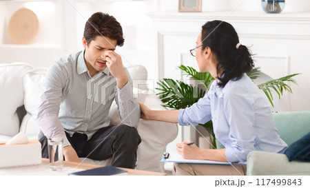 A man sits on a white couch, visibly distressed, holding his forehead while a therapist provides support. The session occurs in a calmly decorated office with modern furnishings and greenery. A man sits on a white couch, visibly distressed, holding his forehead while a therapist provides support. The session occurs in a calmly decorated office with modern furnishings and greenery. 117499843