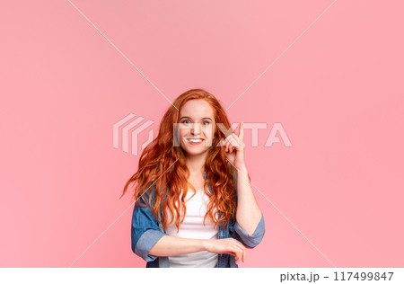 Cheerful young woman with flowing red hair is standing against a pink backdrop, pointing upward with one finger, her expression is friendly, appears to be engaging with someone or presenting an idea. 117499847