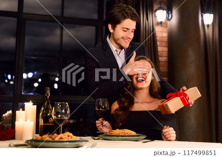 Happy handsome man giving a surprise Valentine's day present to his excited girlfriend, have date at restaurant. Valentine Gift concept 117499851