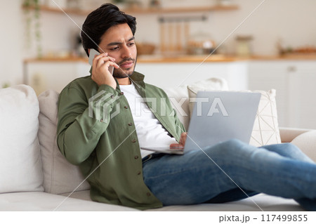 A man is sitting on a white couch and talking on his cell phone. He is wearing a green shirt and blue jeans. He has a laptop on his lap. The man looks focused and serious as he speaks on the phone. 117499855