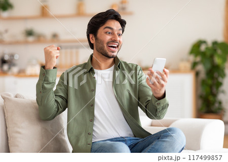 A young man, dressed in a green shirt and jeans, sits on a white couch in his living room. He holding his smartphone in his right hand and celebrating, with his left fist in the air 117499857