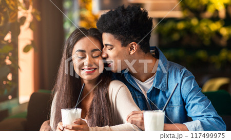 African American young guy and girl are sharing a tender moment as they kiss at a table in a bustling coffee shop. The couple is engrossed in each other, surrounded by coffee cups and a cozy ambiance. 117499975