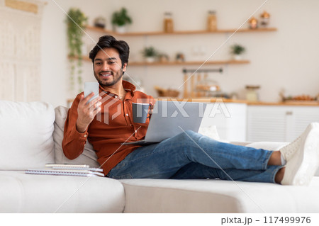 A man is relaxing on a white couch with a laptop on his lap and a smartphone in his hand. He is smiling and looking at his phone. There is a cup of coffee in his other hand 117499976