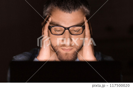 A man wearing glasses concentrates intensely while working on his laptop late at night. His hands are on his head, indicating deep thought or stress. A man wearing glasses concentrates intensely while working on his laptop late at night. His hands are on his head, indicating deep thought or stress. 117500020