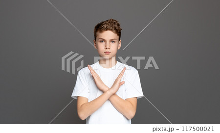 Teenager with arms crossed in an X shape, showcasing refusal or denial, isolated on a grey background 117500021