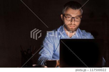 A man wearing glasses is intensely focused while working on his laptop in a dimly lit office environment during late-night hours. He appears to be deeply engaged in a task 117500022