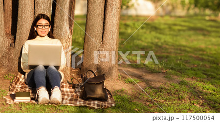 Asian woman is sitting on a blanket outdoors, focused on her laptop. She appears to be working or browsing the internet while enjoying the fresh air. 117500054