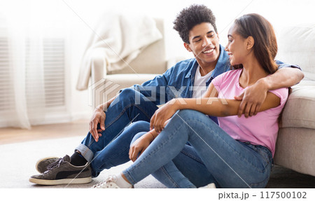 African American teen couple sitting on the floor in a living room. They are both smiling and looking at each other. There is a couch in the background, and the room is decorated in a modern style. African American teen couple sitting on the floor in a living room. They are both smiling and looking at each other. There is a couch in the background, and the room is decorated in a modern style. 117500102