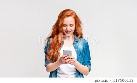 A woman with vibrant red hair is intensely focused on her cell phone screen. She appears engrossed in whatever she is viewing, holding the device with both hands 117500112