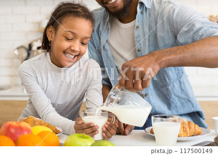 A smiling African American young girl watches as her father pours milk into a glass in a brightly lit kitchen A smiling African American young girl watches as her father pours milk into a glass in a brightly lit kitchen 117500149
