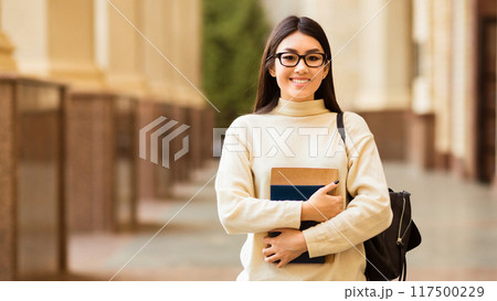 Asian young woman stands confidently on a university campus during the day, smiling while holding a couple of books close to her chest. She is wearing glasses Asian young woman stands confidently on a university campus during the day, smiling while holding a couple of books close to her chest. She is wearing glasses 117500229