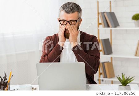 A middle aged man wearing glasses and a sweater sits at a desk in his home office. He is rubbing his eyes in frustration with an open laptop in front of him. Books and office supplies are arranged 117500250
